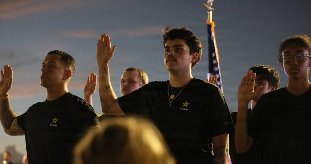 A group of individuals wearing black U.S. Army branded t-shirts are raising their right hands, appearing to take an oath or pledge. An American flag is visible in the background. The scene appears to take place outdoors at dusk or dawn.