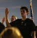 A group of individuals wearing black U.S. Army branded t-shirts are raising their right hands, appearing to take an oath or pledge. An American flag is visible in the background. The scene appears to take place outdoors at dusk or dawn.