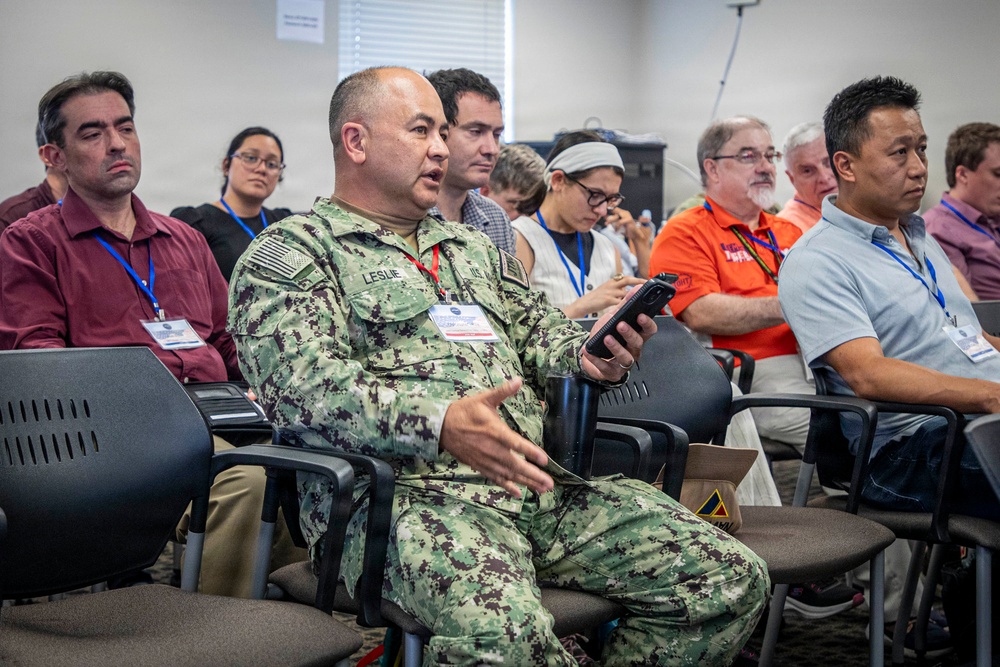 A group of people are seated in rows of chairs at what appears to be a conference or briefing session. A U.S. Navy servicemember in NWU Type III camouflage uniform, identified by the name tape 'LESLIE,' is prominently featured in the foreground holding a device and appearing to speak or ask a question. Attendees are wearing blue lanyards with name badges, suggesting a formal organized event.