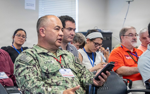A group of people are seated in rows of chairs at what appears to be a conference or briefing session. A U.S. Navy servicemember in NWU Type III camouflage uniform, identified by the name tape 'LESLIE,' is prominently featured in the foreground holding a device and appearing to speak or ask a question. Attendees are wearing blue lanyards with name badges, suggesting a formal organized event.