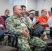 A group of people are seated in rows of chairs at what appears to be a conference or briefing session. A U.S. Navy servicemember in NWU Type III camouflage uniform, identified by the name tape 'LESLIE,' is prominently featured in the foreground holding a device and appearing to speak or ask a question. Attendees are wearing blue lanyards with name badges, suggesting a formal organized event.