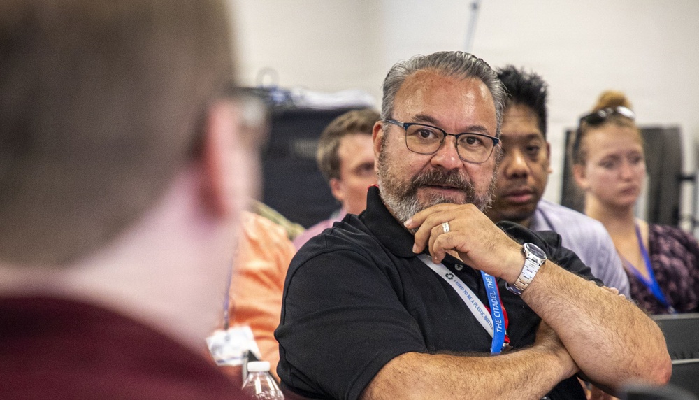 A group of people appear to be attending a seminar, lecture, or meeting. The central figure is a middle-aged man wearing glasses and a black polo shirt with a blue lanyard, listening attentively with his hand raised to his chin in a thoughtful pose. Several other attendees are visible in the background, slightly out of focus.