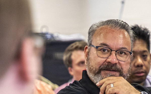 A group of people appear to be attending a seminar, lecture, or meeting. The central figure is a middle-aged man wearing glasses and a black polo shirt with a blue lanyard, listening attentively with his hand raised to his chin in a thoughtful pose. Several other attendees are visible in the background, slightly out of focus.