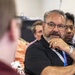A group of people appear to be attending a seminar, lecture, or meeting. The central figure is a middle-aged man wearing glasses and a black polo shirt with a blue lanyard, listening attentively with his hand raised to his chin in a thoughtful pose. Several other attendees are visible in the background, slightly out of focus.