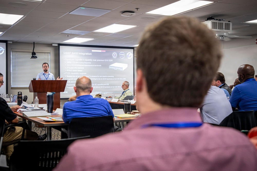 A professional technical briefing or conference is taking place in a modern meeting room. A presenter stands at a podium delivering a presentation about 'USRP X410 Integration,' visible on a large projection screen. Multiple attendees are seated at tables facing the presenter.