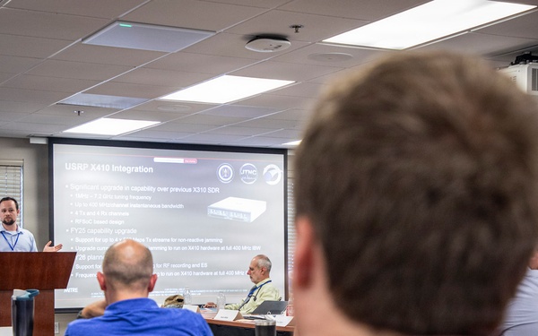 A professional technical briefing or conference is taking place in a modern meeting room. A presenter stands at a podium delivering a presentation about 'USRP X410 Integration,' visible on a large projection screen. Multiple attendees are seated at tables facing the presenter.
