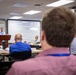 A professional technical briefing or conference is taking place in a modern meeting room. A presenter stands at a podium delivering a presentation about 'USRP X410 Integration,' visible on a large projection screen. Multiple attendees are seated at tables facing the presenter.