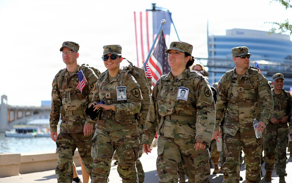 A group of U.S. Army soldiers in MultiCam (OCP) camouflage uniforms are walking together in what appears to be a commemorative ruck march or memorial event near a waterfront. Several soldiers are carrying small American flags and wearing photo badges on their uniforms, suggesting this is a tribute or memorial walk honoring fallen service members. The setting appears to be an urban waterfront area with a large American flag visible in the background.