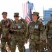 A group of U.S. Army soldiers in MultiCam (OCP) camouflage uniforms are walking together in what appears to be a commemorative ruck march or memorial event near a waterfront. Several soldiers are carrying small American flags and wearing photo badges on their uniforms, suggesting this is a tribute or memorial walk honoring fallen service members. The setting appears to be an urban waterfront area with a large American flag visible in the background.