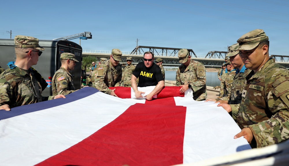 A group of U.S. Army soldiers in camouflage uniforms, along with one individual in a black 'ARMY' t-shirt, are carefully folding a large American flag. The scene takes place outdoors near a waterway with a bridge visible in the background. This appears to be a formal or ceremonial flag-folding procedure.