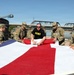 A group of U.S. Army soldiers in camouflage uniforms, along with one individual in a black 'ARMY' t-shirt, are carefully folding a large American flag. The scene takes place outdoors near a waterway with a bridge visible in the background. This appears to be a formal or ceremonial flag-folding procedure.