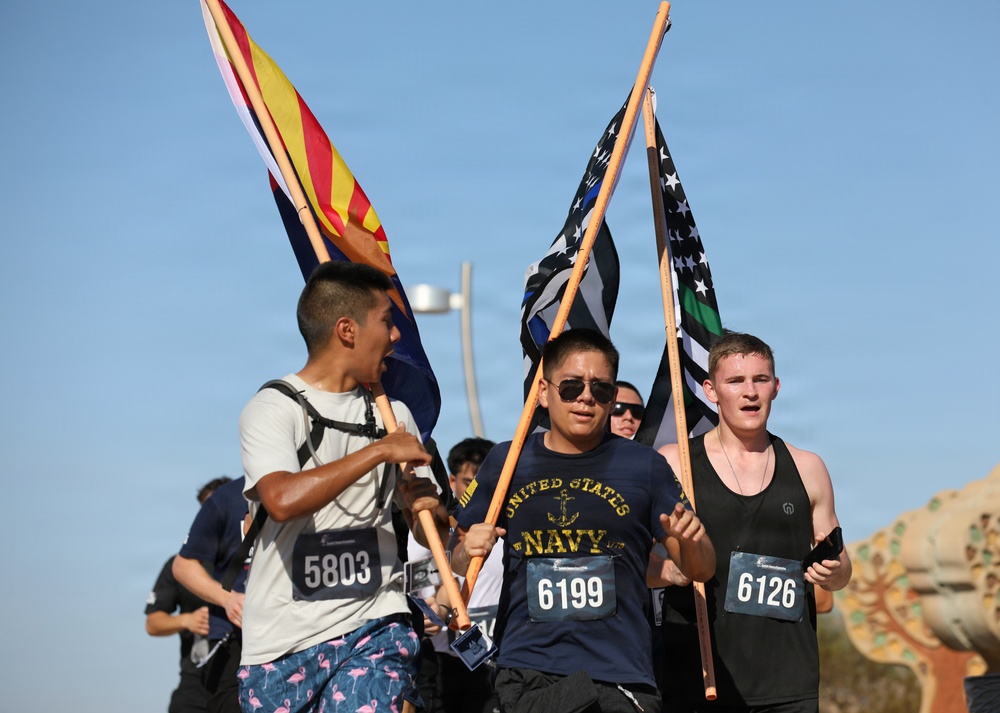Three runners participate in a race event, carrying flags while wearing numbered bibs. The central runner wears a United States Navy t-shirt and sunglasses, carrying what appears to be a Thin Blue Line / Thin Green Line flag, while the runner on the left carries what appears to be the Arizona state flag. The event appears to be an outdoor race held in a sunny, arid environment.