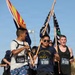 Three runners participate in a race event, carrying flags while wearing numbered bibs. The central runner wears a United States Navy t-shirt and sunglasses, carrying what appears to be a Thin Blue Line / Thin Green Line flag, while the runner on the left carries what appears to be the Arizona state flag. The event appears to be an outdoor race held in a sunny, arid environment.