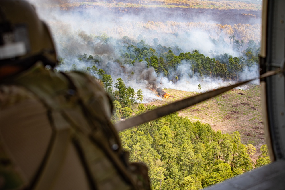 An aerial view taken from inside a military helicopter showing a fire burning in a cleared field surrounded by pine forest. A soldier in camouflage uniform and helmet is visible in the foreground observing the scene below. Multiple columns of smoke rise from active flames in what appears to be a controlled burn or strike zone.