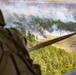 An aerial view taken from inside a military helicopter showing a fire burning in a cleared field surrounded by pine forest. A soldier in camouflage uniform and helmet is visible in the foreground observing the scene below. Multiple columns of smoke rise from active flames in what appears to be a controlled burn or strike zone.