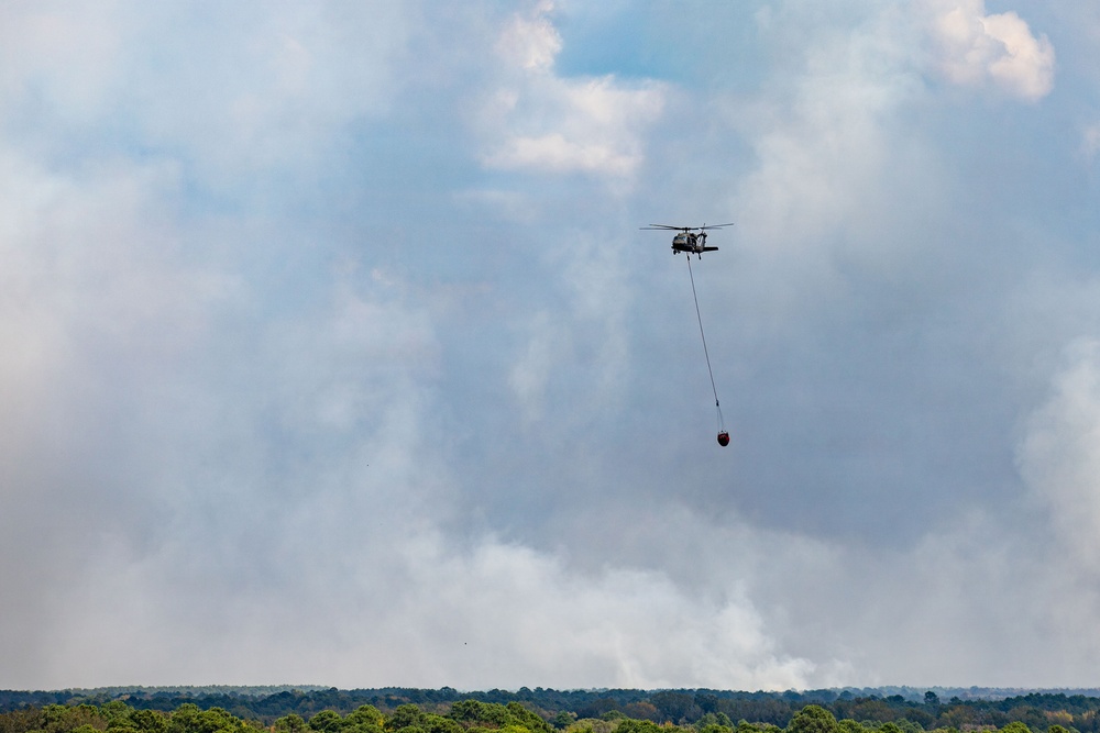 A military-style helicopter is airborne carrying a suspended firefighting water bucket (Bambi bucket) beneath it via a long cable. Dense white and gray smoke billows in the background above a treeline, indicating an active wildfire below. The helicopter appears to be conducting aerial firefighting operations over a forested area.