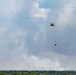 A military-style helicopter is airborne carrying a suspended firefighting water bucket (Bambi bucket) beneath it via a long cable. Dense white and gray smoke billows in the background above a treeline, indicating an active wildfire below. The helicopter appears to be conducting aerial firefighting operations over a forested area.