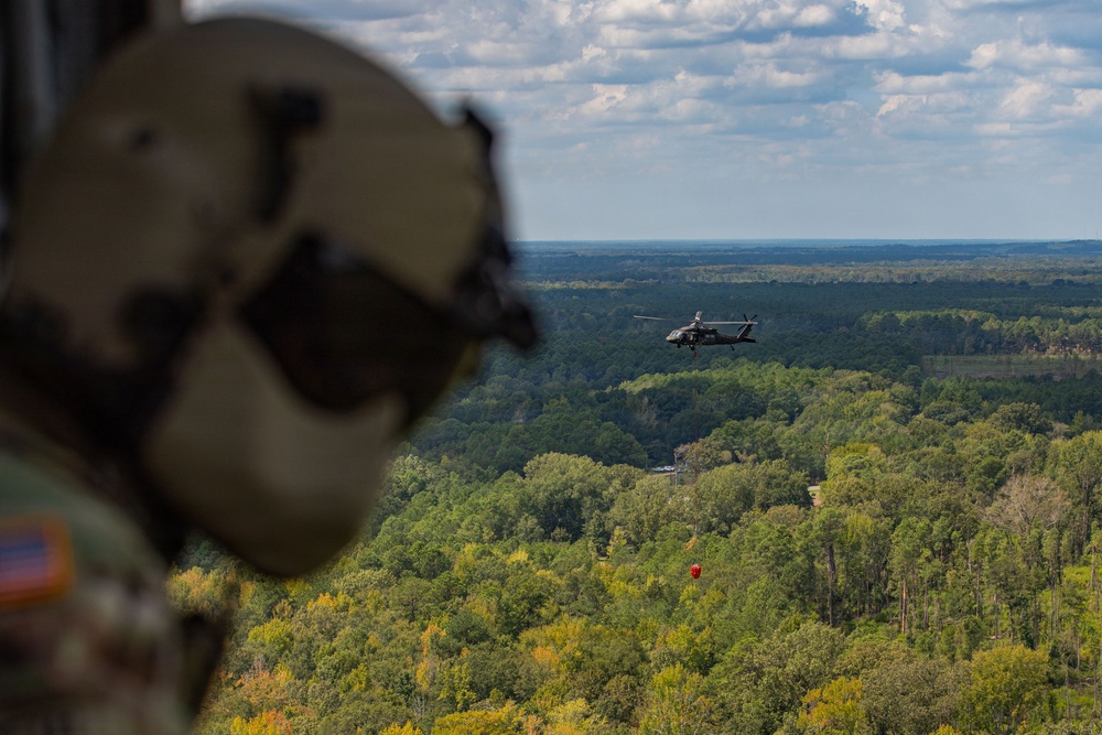 A military aviator wearing a flight helmet and camouflage uniform observes from an aircraft as a helicopter — likely a UH-60 Black Hawk — flies over a dense forested landscape. The distant helicopter appears to be carrying a suspended load, possibly a water bucket or sling-load, indicating a utility or firefighting operation. The scene is captured from an aerial vantage point with a wide, tree-covered terrain and partly cloudy sky in the background.