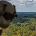 A military aviator wearing a flight helmet and camouflage uniform observes from an aircraft as a helicopter — likely a UH-60 Black Hawk — flies over a dense forested landscape. The distant helicopter appears to be carrying a suspended load, possibly a water bucket or sling-load, indicating a utility or firefighting operation. The scene is captured from an aerial vantage point with a wide, tree-covered terrain and partly cloudy sky in the background.