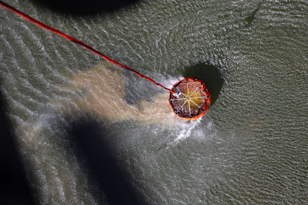 An aerial photograph shows a circular orange flotation device or containment buoy being towed across choppy, greenish water by a red rope or cable. The device appears to be in motion, creating a visible wake and disturbed water trail behind it. The image is taken from directly above, likely from a helicopter or drone.