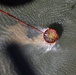 An aerial photograph shows a circular orange flotation device or containment buoy being towed across choppy, greenish water by a red rope or cable. The device appears to be in motion, creating a visible wake and disturbed water trail behind it. The image is taken from directly above, likely from a helicopter or drone.