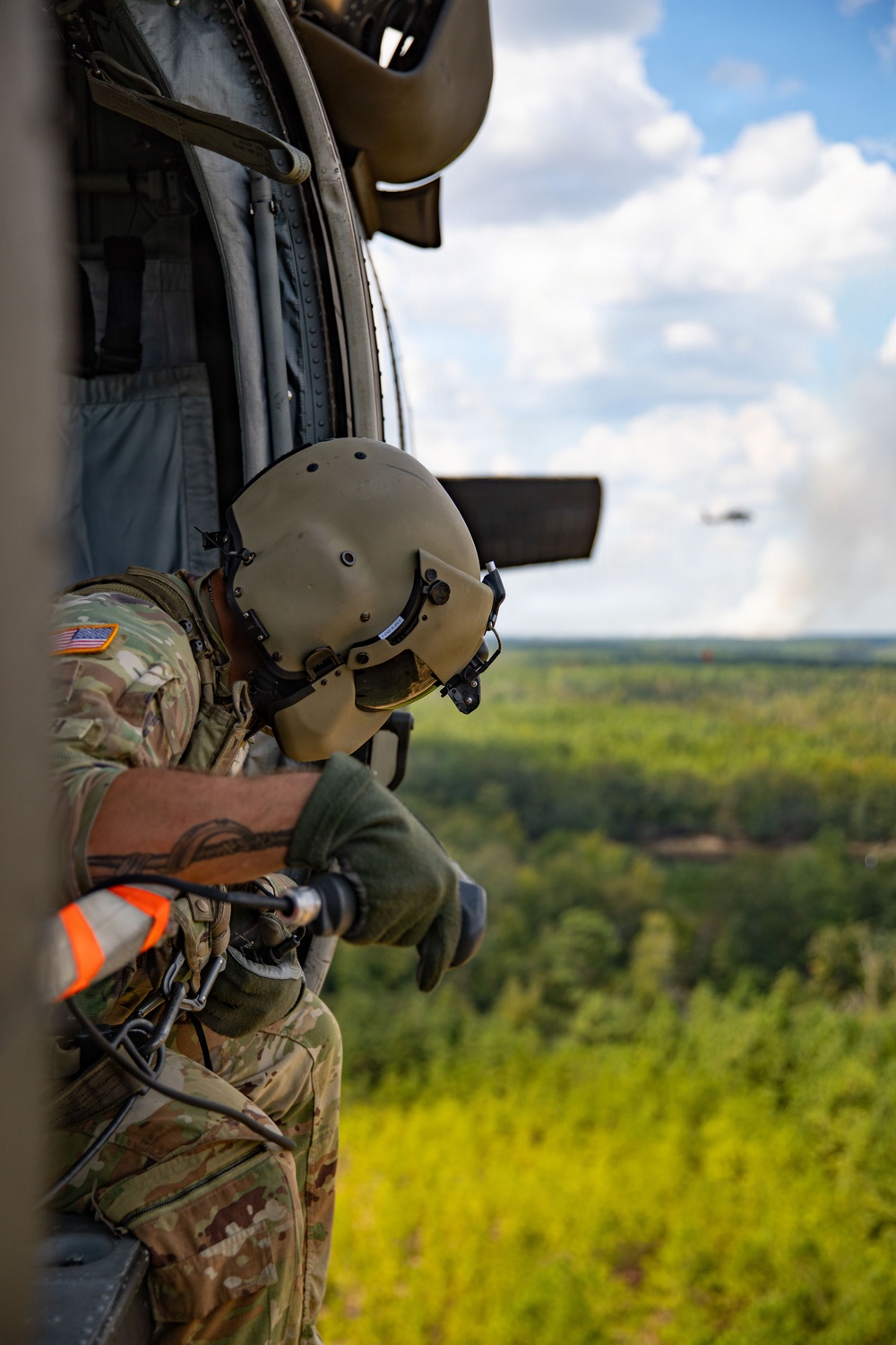 A U.S. Army soldier is leaning out of the open door of a military helicopter in flight, looking downward over a dense green forested landscape. The soldier is wearing OCP (Operational Camouflage Pattern) uniform, a flight/combat helmet with visor, and green tactical gloves, and appears to be tethered to the aircraft via a safety harness with orange high-visibility components. A second helicopter is visible in the background sky.