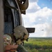 A U.S. Army soldier is leaning out of the open door of a military helicopter in flight, looking downward over a dense green forested landscape. The soldier is wearing OCP (Operational Camouflage Pattern) uniform, a flight/combat helmet with visor, and green tactical gloves, and appears to be tethered to the aircraft via a safety harness with orange high-visibility components. A second helicopter is visible in the background sky.
