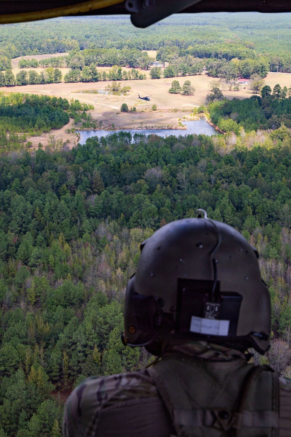 A military helicopter crew member in full flight gear observes another helicopter flying low over a rural landscape from an open door or window of their aircraft. The scene depicts a formation flight or training exercise over mixed terrain of forests, open fields, and small bodies of water. The perspective is from inside one aircraft looking outward toward the second helicopter in the distance.