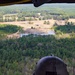 A military helicopter crew member in full flight gear observes another helicopter flying low over a rural landscape from an open door or window of their aircraft. The scene depicts a formation flight or training exercise over mixed terrain of forests, open fields, and small bodies of water. The perspective is from inside one aircraft looking outward toward the second helicopter in the distance.