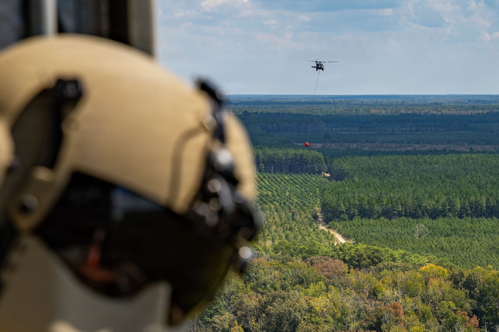 An aerial photograph taken from inside a helicopter showing a crew member in a tan flight helmet observing another helicopter in the distance carrying a red water bucket suspended by a cable. The scene is set over a vast forested landscape with rows of pine trees and a dirt road visible below. This appears to be a firefighting or water-bucket training operation.