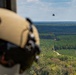 An aerial photograph taken from inside a helicopter showing a crew member in a tan flight helmet observing another helicopter in the distance carrying a red water bucket suspended by a cable. The scene is set over a vast forested landscape with rows of pine trees and a dirt road visible below. This appears to be a firefighting or water-bucket training operation.