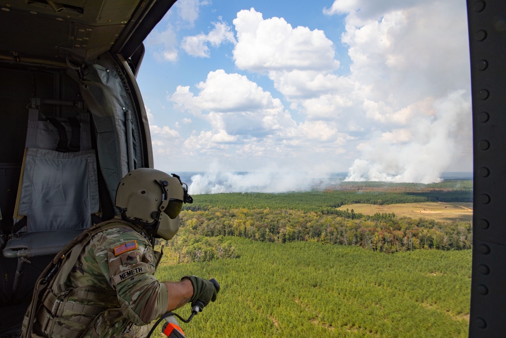 A U.S. Army soldier identified as 'NEMETH' observes a large wildfire from the open door of a military helicopter. The aerial perspective shows extensive smoke columns rising from a forested landscape below. The soldier is wearing full combat gear including a flight helmet and multicam uniform with a U.S. flag patch.