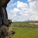 A U.S. Army soldier identified as 'NEMETH' observes a large wildfire from the open door of a military helicopter. The aerial perspective shows extensive smoke columns rising from a forested landscape below. The soldier is wearing full combat gear including a flight helmet and multicam uniform with a U.S. flag patch.