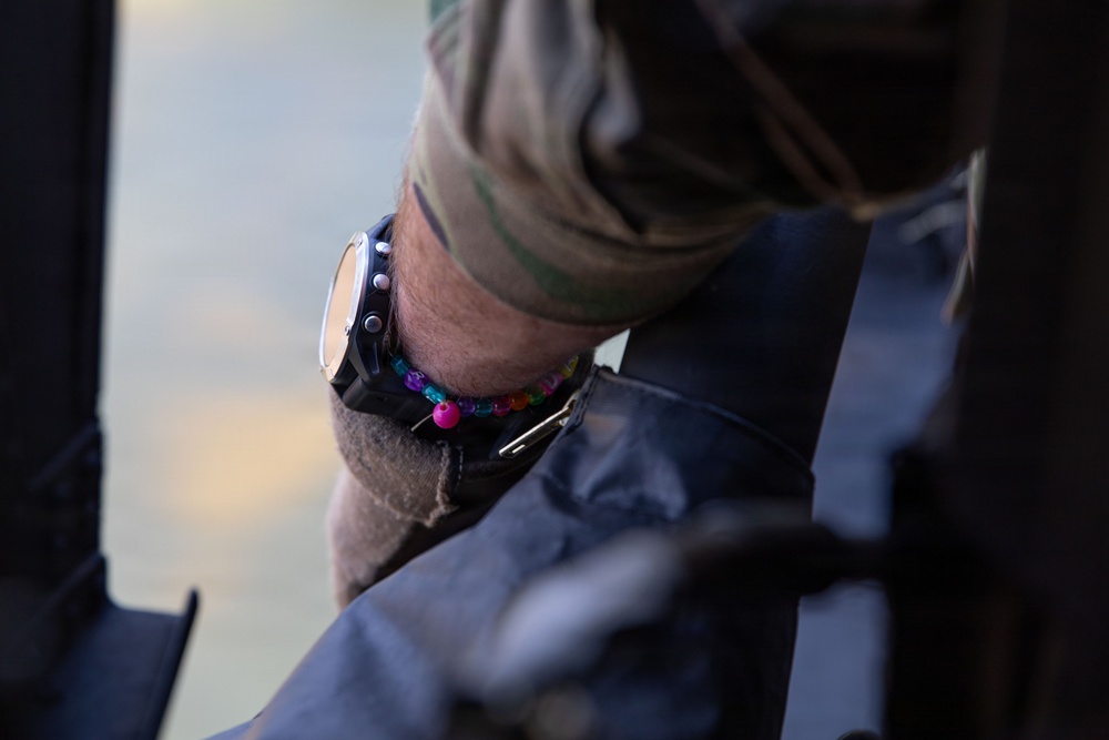 A close-up photograph of a person's wrist wearing a black digital/tactical watch alongside a colorful beaded bracelet with multicolored beads including pink, blue, orange, and green. The person is wearing a camouflage-patterned sleeve, suggesting military or tactical attire. The wrist appears to be resting on or near a dark-colored surface or equipment.