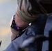 A close-up photograph of a person's wrist wearing a black digital/tactical watch alongside a colorful beaded bracelet with multicolored beads including pink, blue, orange, and green. The person is wearing a camouflage-patterned sleeve, suggesting military or tactical attire. The wrist appears to be resting on or near a dark-colored surface or equipment.