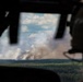 The image is taken from inside an aircraft cockpit, showing a person wearing a flight helmet observing a large smoke plume rising from a forested area in the distance. The smoke column is substantial and billowing upward, consistent with a wildfire or controlled burn. The perspective through the aircraft window frames the scene dramatically against a partly cloudy sky.