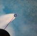 The image captures a small parachute or drogue chute being ejected from what appears to be a cannon or launch device, trailing a red cord amid a plume of white smoke. The perspective is taken from close to the launch apparatus, looking upward and outward. Trees are visible in the background through the haze of smoke.