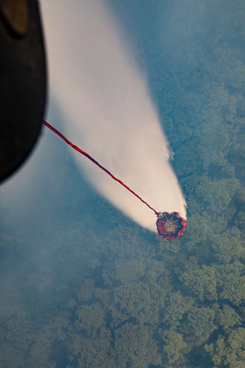 An aerial photograph taken from an aircraft showing a firefighting helicopter or fixed-wing aircraft deploying water or fire retardant from a suspended bucket (Bambi bucket) over a forested area. A red and blue tether connects the aircraft to the red circular bucket, which is releasing a wide cone-shaped spray of white liquid downward. The terrain below appears to be dense green forest canopy viewed from significant altitude.