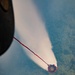 An aerial photograph taken from an aircraft showing a firefighting helicopter or fixed-wing aircraft deploying water or fire retardant from a suspended bucket (Bambi bucket) over a forested area. A red and blue tether connects the aircraft to the red circular bucket, which is releasing a wide cone-shaped spray of white liquid downward. The terrain below appears to be dense green forest canopy viewed from significant altitude.
