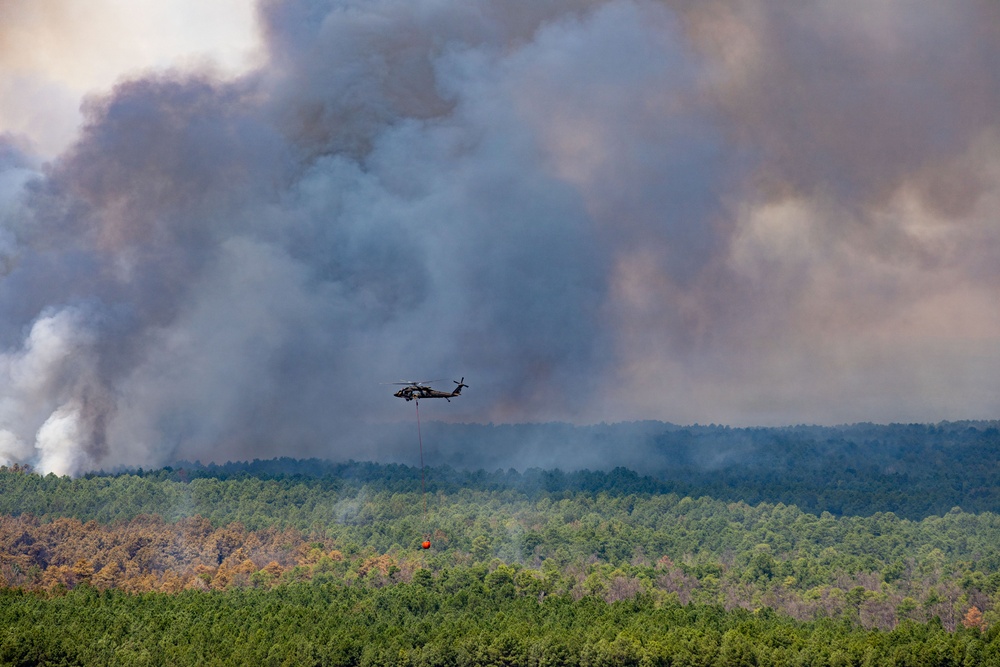 A military-style helicopter, likely a UH-60 Black Hawk, is conducting aerial firefighting operations over a densely forested area. The aircraft is carrying a suspended orange water/retardant bucket (Bambi bucket) beneath it. Massive columns of gray and white smoke billow in the background, indicating a large active wildfire in the forest.