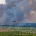 A military-style helicopter, likely a UH-60 Black Hawk, is conducting aerial firefighting operations over a densely forested area. The aircraft is carrying a suspended orange water/retardant bucket (Bambi bucket) beneath it. Massive columns of gray and white smoke billow in the background, indicating a large active wildfire in the forest.