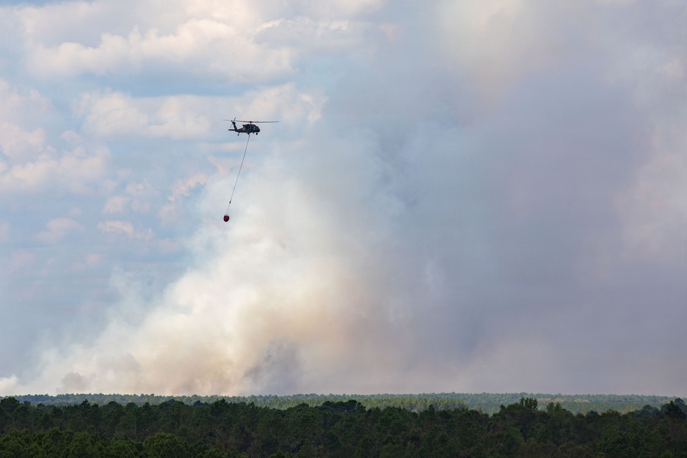 A helicopter is conducting aerial firefighting operations over a forested area, carrying a suspended water/retardant bucket on a long line beneath it. Large plumes of white and gray smoke rise from the tree line below, indicating an active wildfire. The helicopter appears to be maneuvering toward or away from the fire zone.