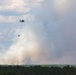 A helicopter is conducting aerial firefighting operations over a forested area, carrying a suspended water/retardant bucket on a long line beneath it. Large plumes of white and gray smoke rise from the tree line below, indicating an active wildfire. The helicopter appears to be maneuvering toward or away from the fire zone.