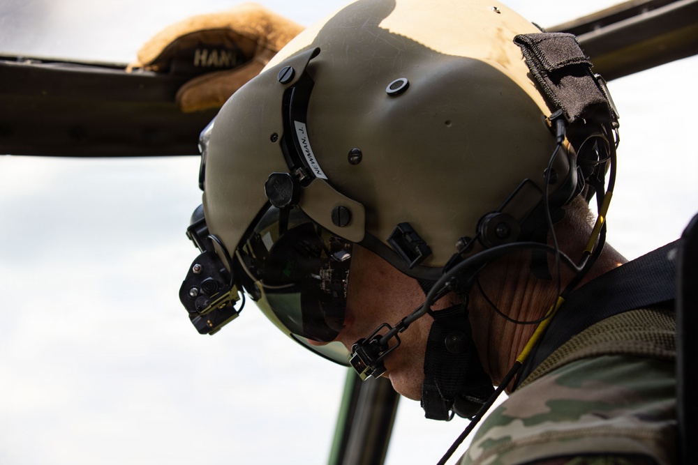 A military aviator or crew member is photographed in close-up profile view, wearing a tactical flight helmet with integrated communications equipment and night vision goggle mounts. The individual is dressed in camouflage uniform and appears to be leaning out of or inspecting a military aircraft. A name label reading 'NEWMAN' is visible on the helmet.