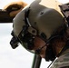 A military aviator or crew member is photographed in close-up profile view, wearing a tactical flight helmet with integrated communications equipment and night vision goggle mounts. The individual is dressed in camouflage uniform and appears to be leaning out of or inspecting a military aircraft. A name label reading 'NEWMAN' is visible on the helmet.