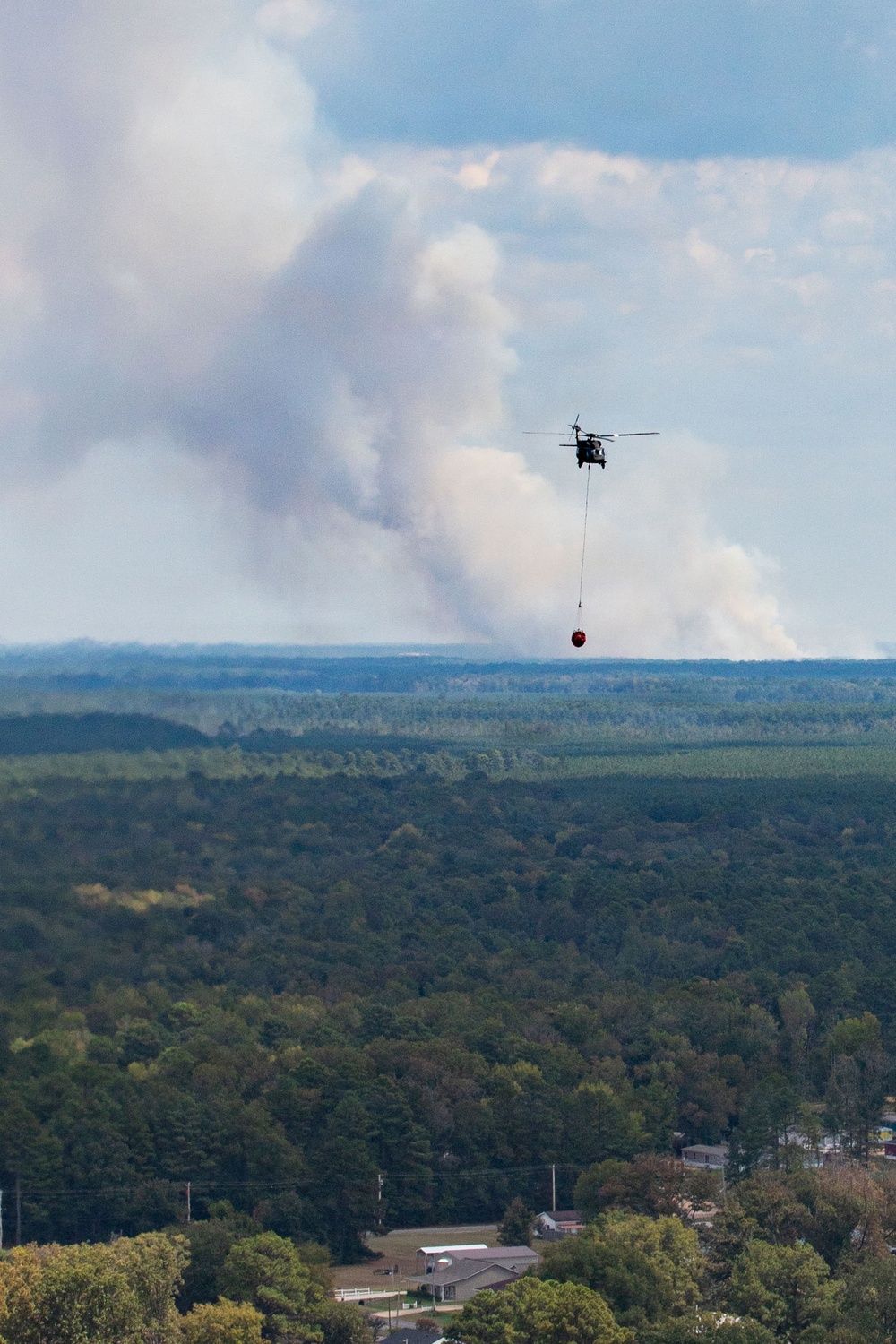 A helicopter is conducting aerial firefighting operations, carrying a red water/retardant bucket suspended by a long line beneath it. Large columns of white and gray smoke rise from a wildfire burning in a forested area in the background. The scene is photographed from an elevated vantage point overlooking a rural/suburban landscape with dense tree cover and scattered buildings.