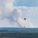 A helicopter is conducting aerial firefighting operations, carrying a red water/retardant bucket suspended by a long line beneath it. Large columns of white and gray smoke rise from a wildfire burning in a forested area in the background. The scene is photographed from an elevated vantage point overlooking a rural/suburban landscape with dense tree cover and scattered buildings.