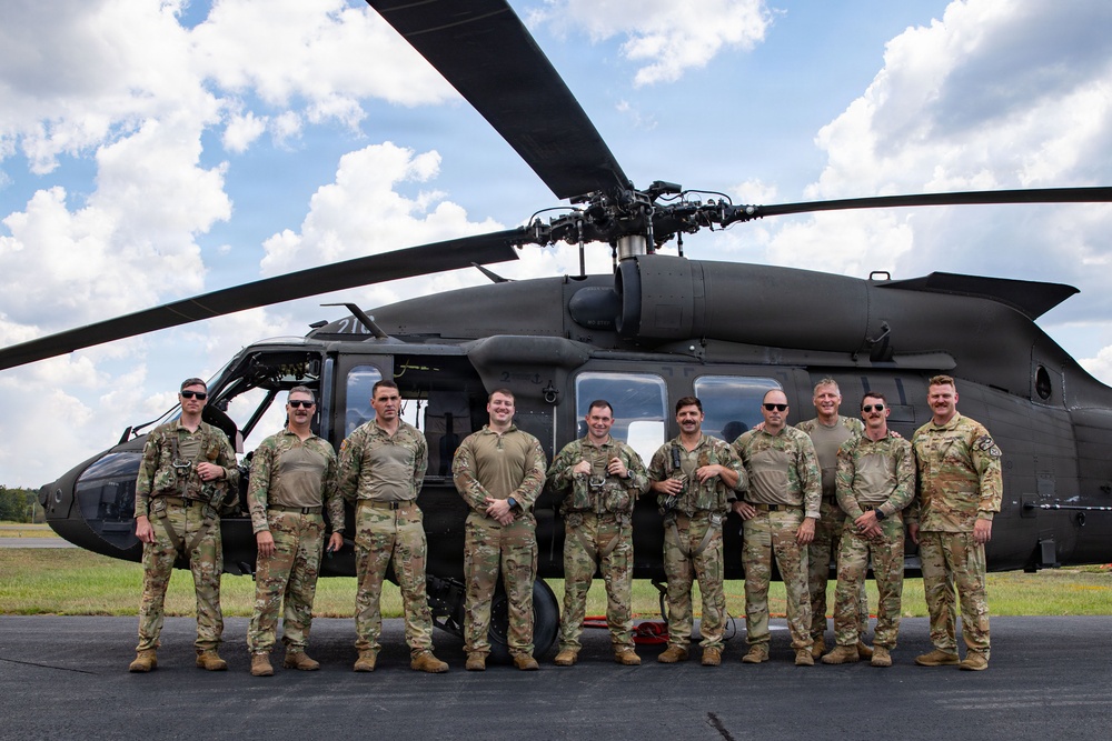 A group of approximately ten military personnel in camouflage uniforms pose for a group photo in front of a UH-60 Black Hawk helicopter on a tarmac. The aircraft is painted in standard military olive/dark green and appears to be parked on a paved surface with a grassy field visible in the background. The image appears to be a formal unit or crew photograph taken in good daylight conditions.