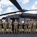 A group of approximately ten military personnel in camouflage uniforms pose for a group photo in front of a UH-60 Black Hawk helicopter on a tarmac. The aircraft is painted in standard military olive/dark green and appears to be parked on a paved surface with a grassy field visible in the background. The image appears to be a formal unit or crew photograph taken in good daylight conditions.