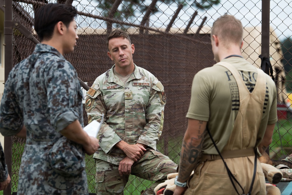 Three military personnel are engaged in a discussion outdoors near a chain-link fence. The central figure wears a US Air Force multicam uniform with the name tape 'MEANEY' and rank insignia visible. A person in what appears to be a foreign military uniform (blue digital camouflage) stands to the left, while a third individual in tan tactical gear with 'POLICE' visible on the back stands to the right.